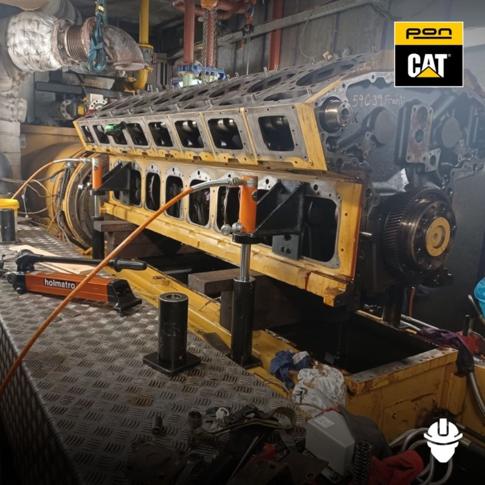 Holmatro cylinder donkraft løfter en tung motorblok i et værksted, omgivet af værktøj og rør.