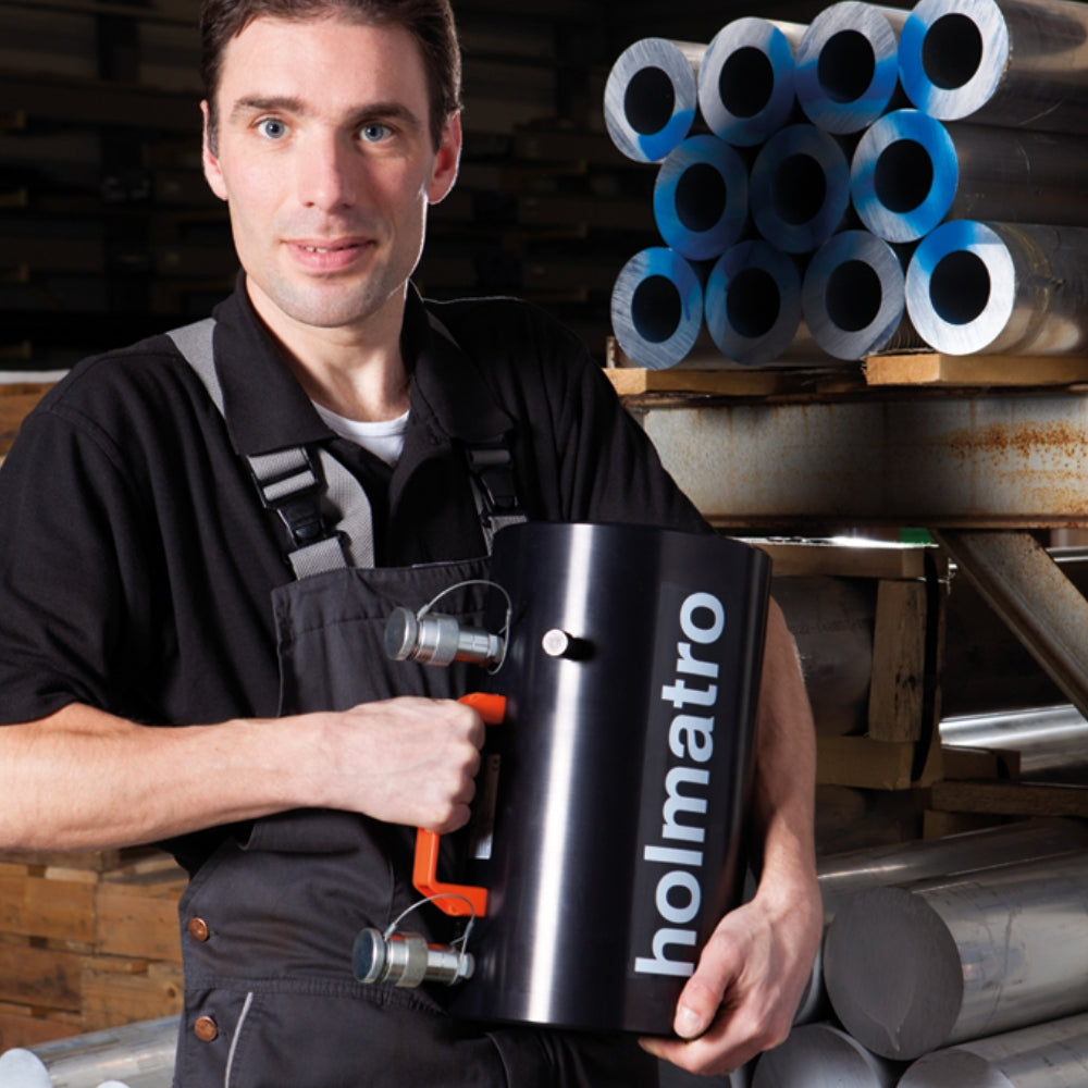 Person holder en sort Holmatro cylinder med orange håndtag, omgivet af metalrør i et værksted.