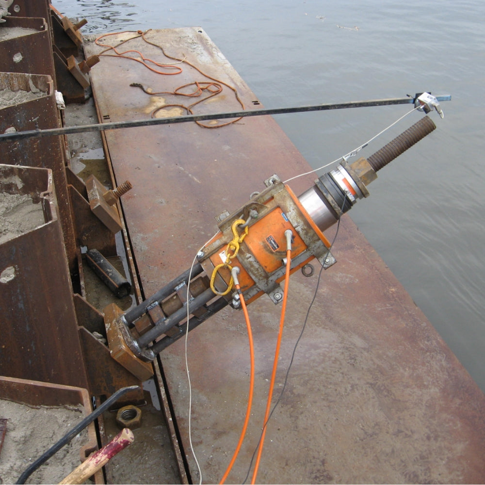 Orange hydraulisk donkraft på rusten metaloverflade ved vand, med tilsluttede ledninger.