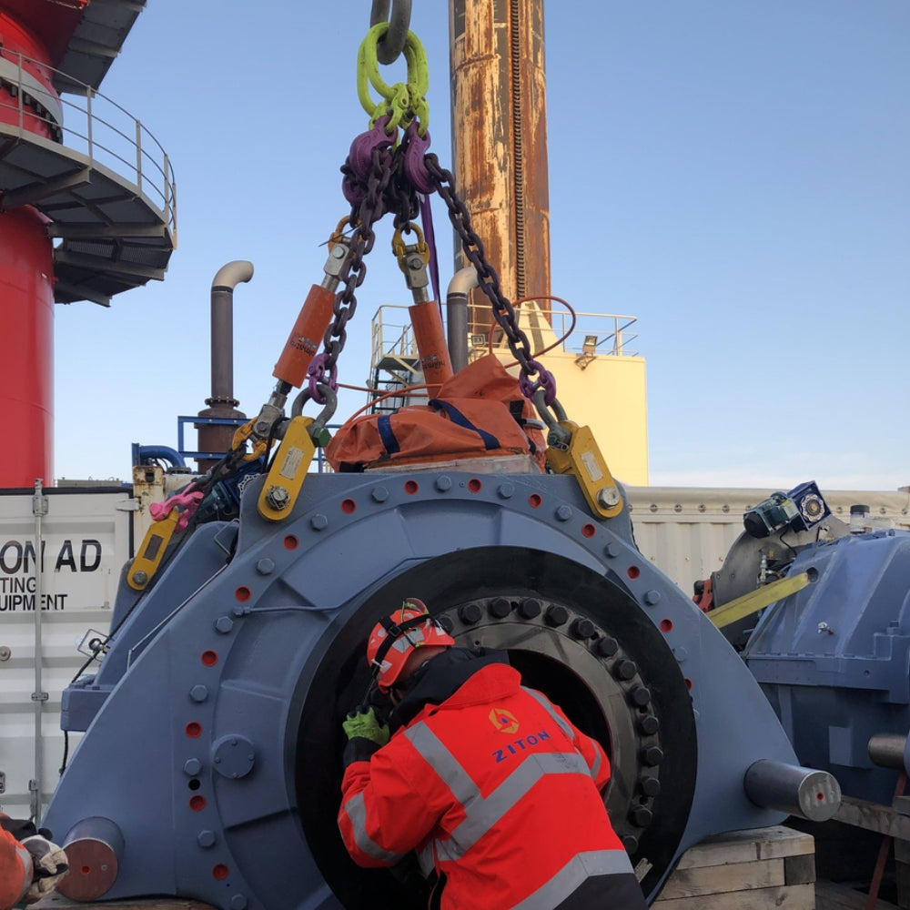 Stor hydraulisk komponent med person i sikkerhedstøj. Krane og udstyr i baggrunden.