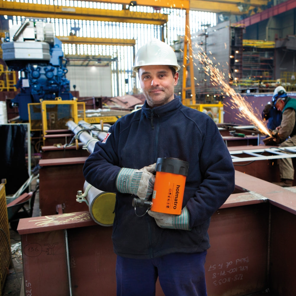 Arbejder i hjelm holder Holmatro cylinder i fabrik. Bagved ses gnister fra en anden arbejder.