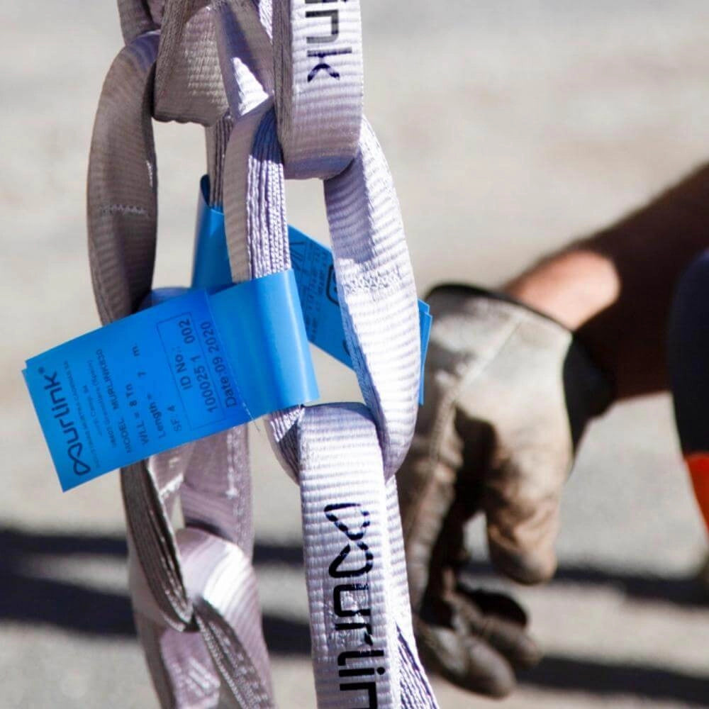 Grå tekstilsløjfer med "Murlink" og blå mærkat. En handsket hånd ses i baggrunden.