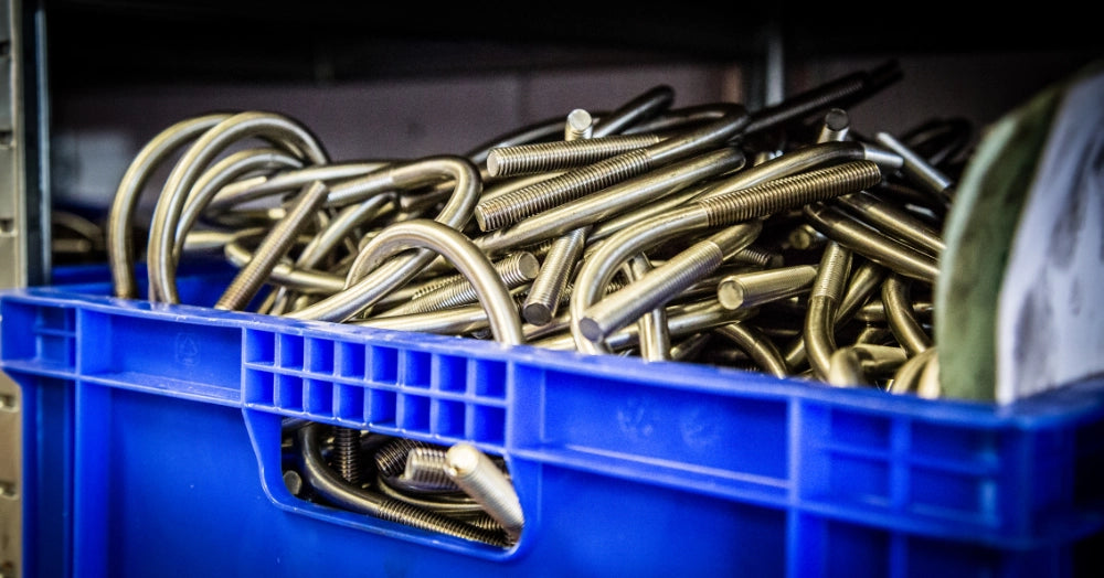 Blå kasse fyldt med metalliske U-bøjler med gevind i begge ender, stablet tæt sammen.