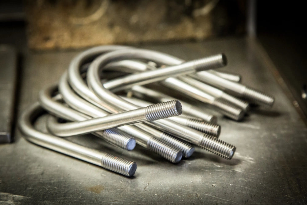 Flere U-bøjler med gevindender og møtrikker, ligger i en bunke på en metaloverflade.